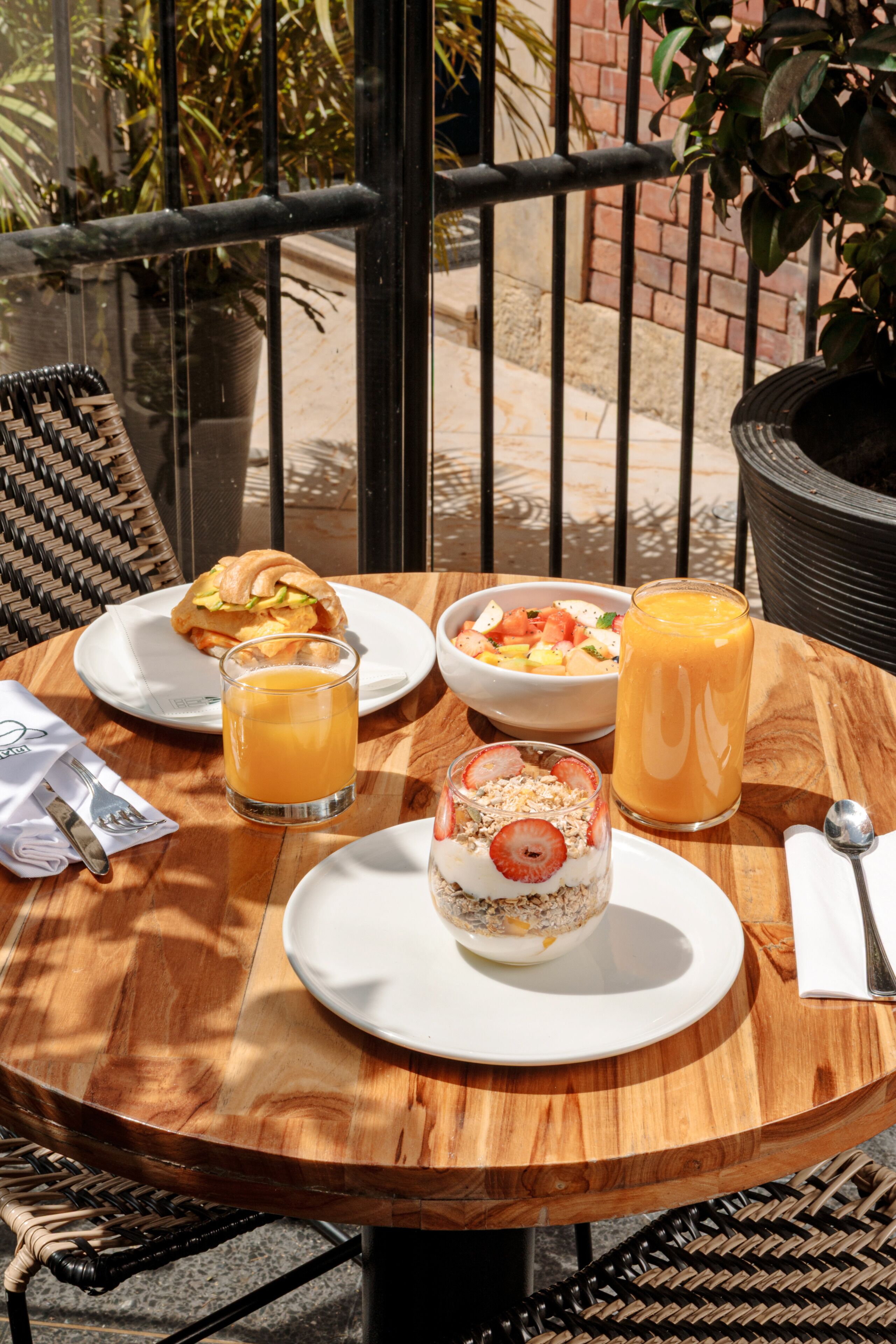 Uma mesa de madeira redonda com itens de café da manhã, incluindo um sanduíche, uma tigela de frutas, um smoothie e sobremesa.
