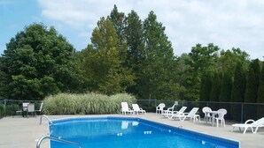 Una piscina rectangular con una escalera metálica, rodeada de sillas de descanso blancas y un césped con plantas altas.