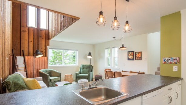 A kitchen with a granite countertop, stainless steel sink, and pendant lighting. There is a green armchair and a wooden dining table with chairs.
