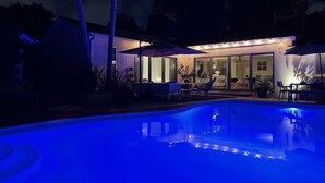 A swimming pool with a blue light, a covered patio area with chairs and umbrellas, and a house in the background. - Ultimate Summer Vacation Home (Los Angeles)