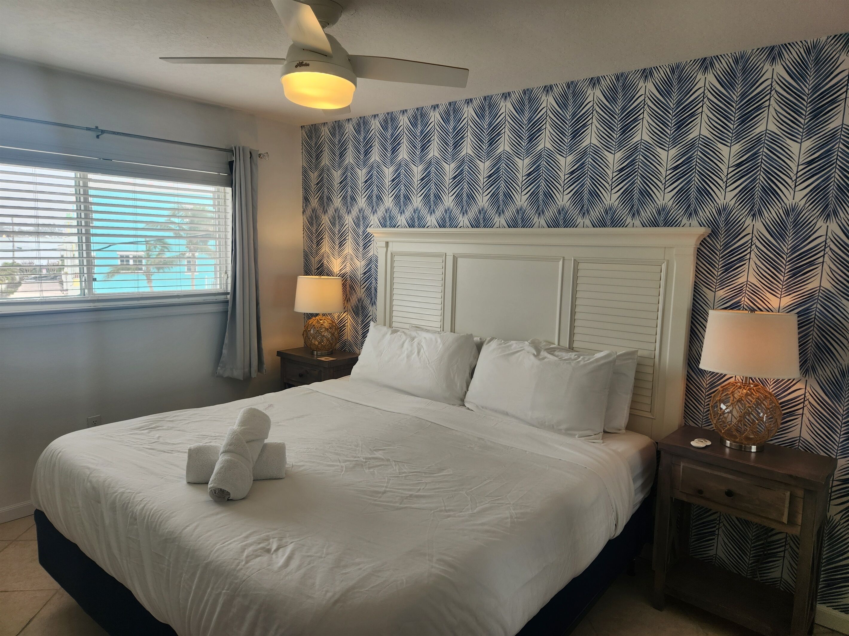 A neatly made bed with white linens and a folded towel, a ceiling fan, two bedside lamps, and a patterned wall.