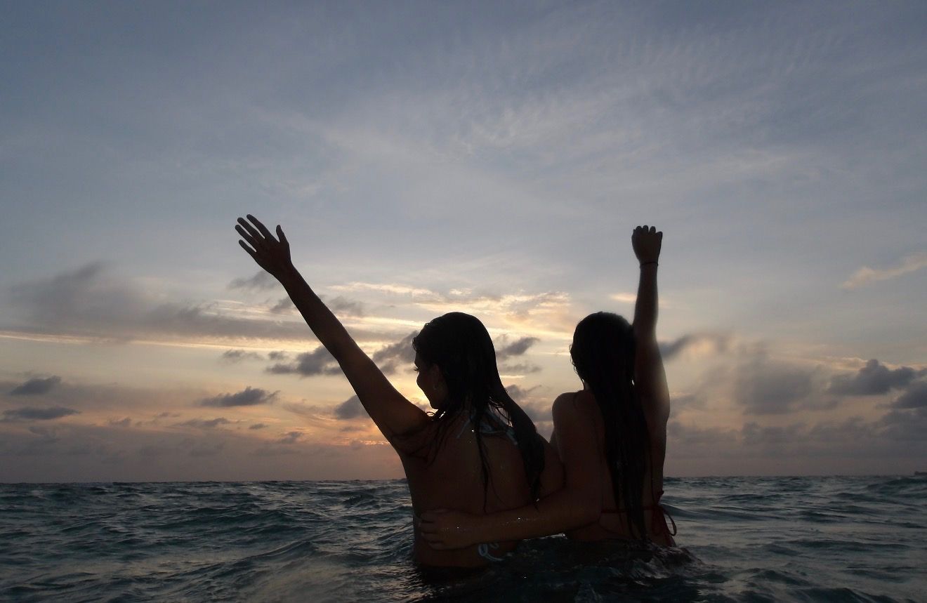 Two people in the ocean with their arms raised against a sunset sky.