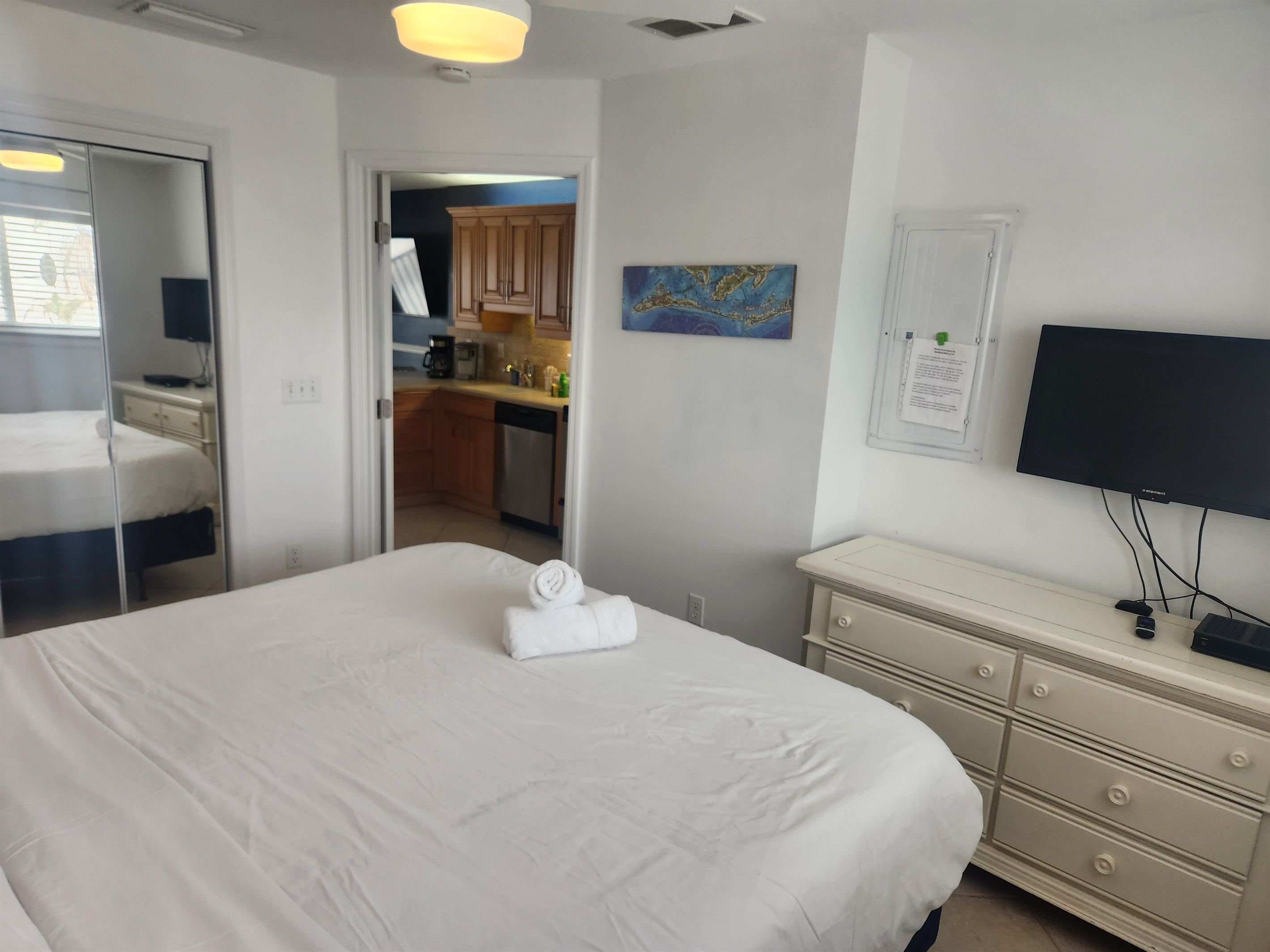 Ein Hotelzimmer mit Bett, Schminktisch, Fernseher und einer sichtbaren Küchenzeile durch eine offene Tür.