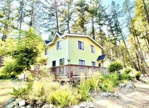 Una casa de dos pisos con un porche de madera, rodeada de un bosque denso y terreno rocoso.
