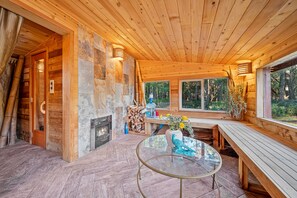 Interior de una cabaña de madera con una mesa de vidrio, bancos, chimenea y una pared de piedra.