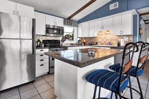 A modern kitchen with white cabinets, a stainless steel refrigerator, a built-in oven, and a granite island with bar stools.