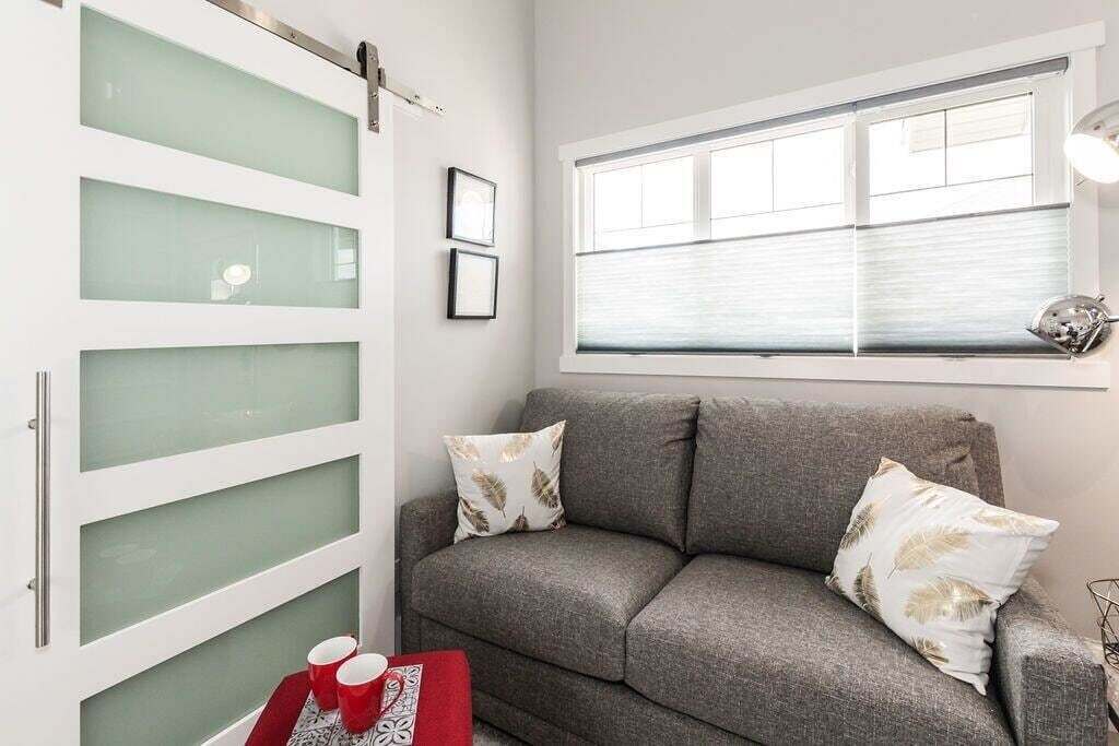 A modern living room with a grey sofa, two decorative pillows, a red tray with cups, and a window with blinds.