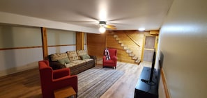 Une pièce avec un escalier en bois, un canapé, un fauteuil rouge, une télévision et un ventilateur de plafond.