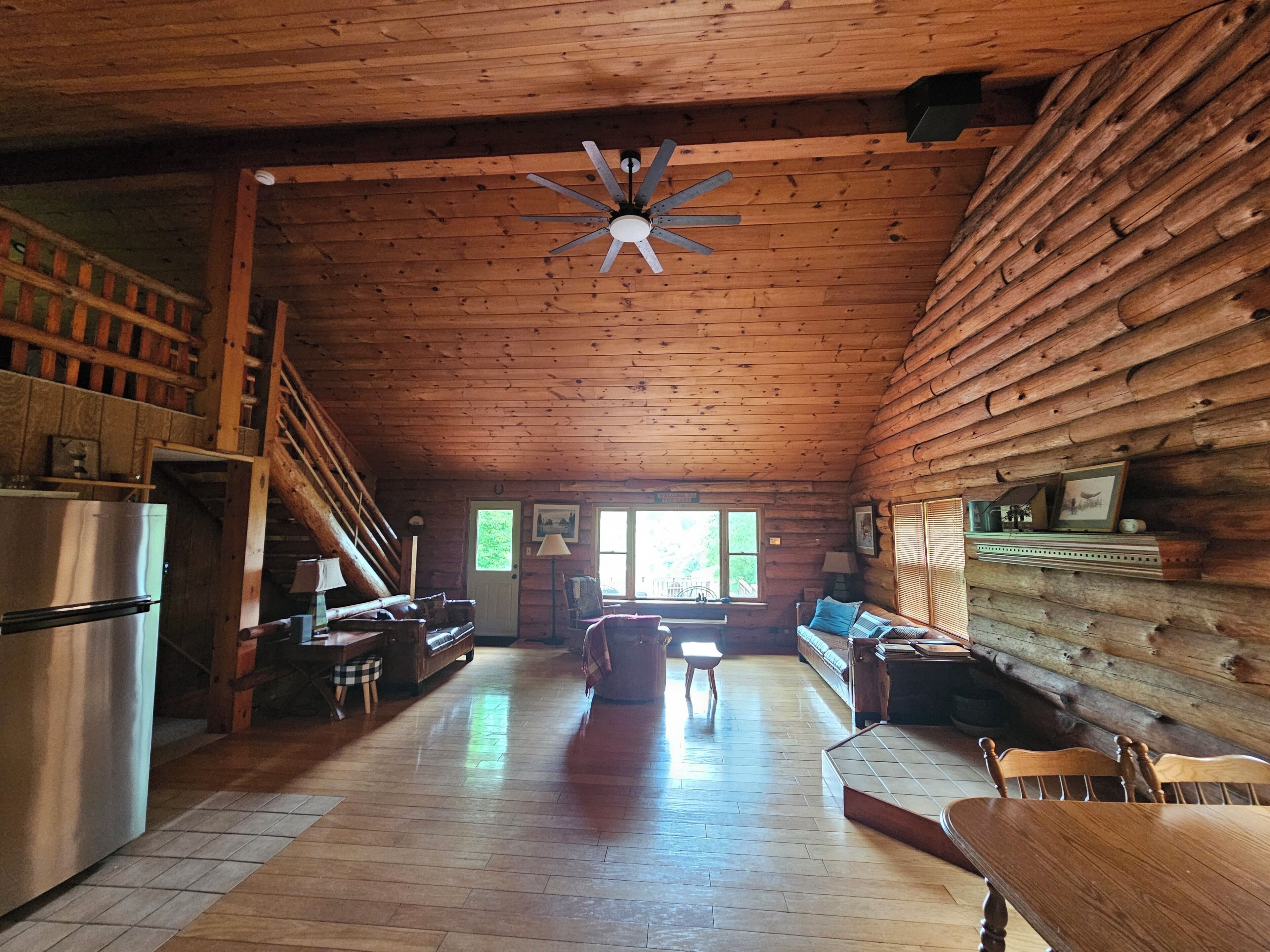 A log cabin interior with a wooden ceiling, a central ceiling fan, a kitchen area with a refrigerator, a dining table, and a staircase.