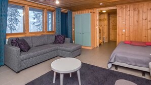 A cozy cabin interior with a sofa, a bed, a round table, and a view of snowy trees through large windows.