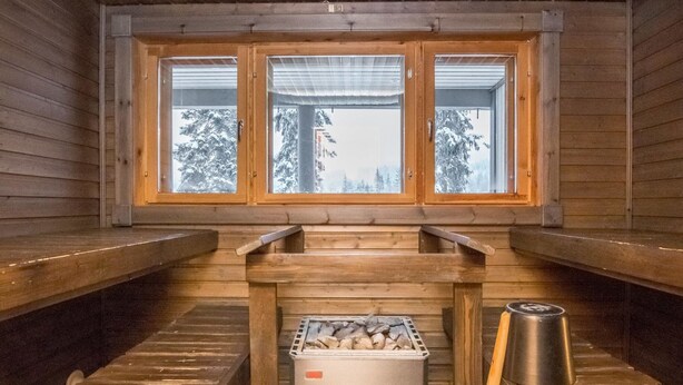A sauna room with wooden benches, a fire pit, and a window with a view of snow-covered trees.