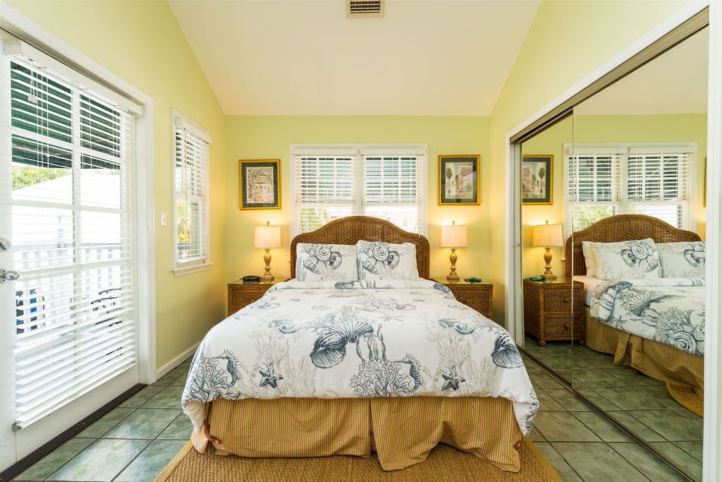 A bedroom with a bed, two bedside tables, two lamps, a mirror, and a window with blinds.