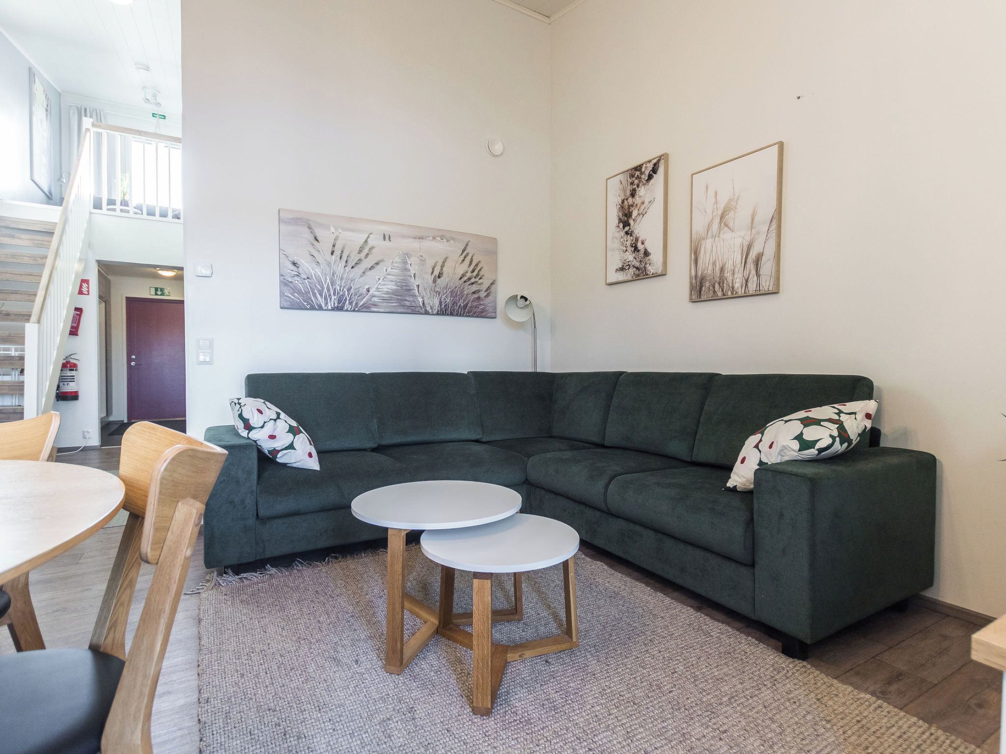 Una sala moderna con un sofá seccional verde, dos mesitas redondas blancas, una mesa de comedor de madera con sillas y una escalera que sube a un nivel superior.