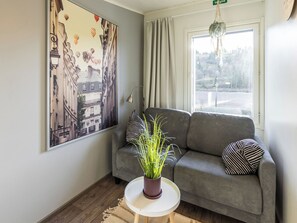 A room with a grey sofa, striped pillows, a potted plant on a small table, a large framed picture on the wall, and a window with curtains.