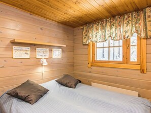Habitación de cabaña de madera con cama, dos almohadas, mesita de noche, lámpara y ventana con cortinas.