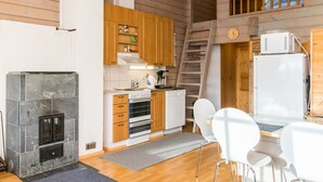 A compact kitchen with a stone fireplace, wooden cabinets, and a small dining area with white chairs. - Gasthalla b paritalo by Interhome (Hyrynsalmi)