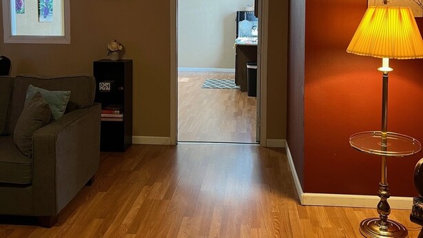 Un salón con suelo de madera, un sofá, una lámpara y una puerta que da a otra habitación.