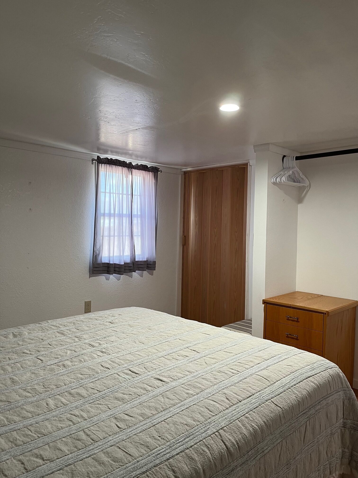 A bedroom with a bed, a wooden wardrobe, a window with curtains, and a hanging clothes rack.