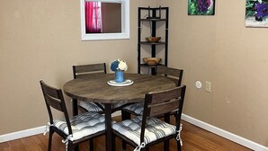 Un comedor con una mesa redonda de madera, seis sillas con cojines a rayas, un florero azul con flores, una ventana con cortinas rosadas y dos estantes montados en la pared con objetos decorativos.