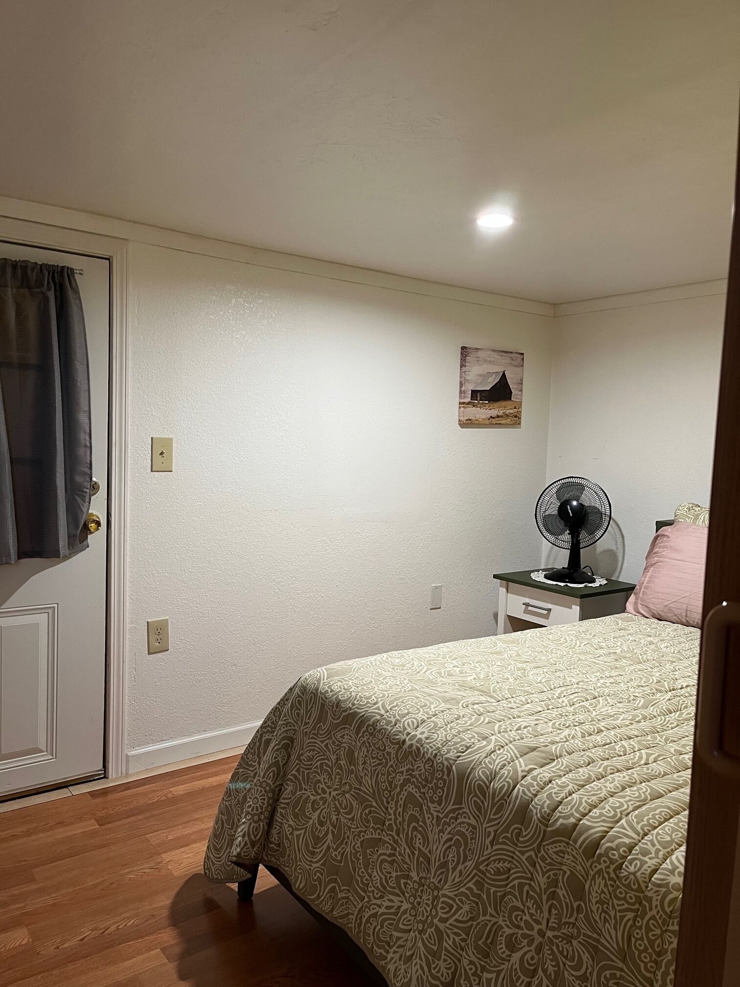 A bedroom with a bed, a nightstand with a fan, a door with a curtain, and a wall-mounted picture.
