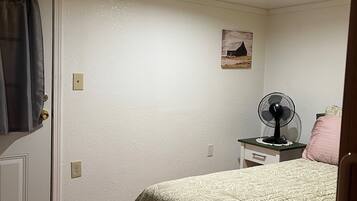 A bedroom with a bed, a nightstand with a fan, a door with a curtain, and a wall-mounted picture.