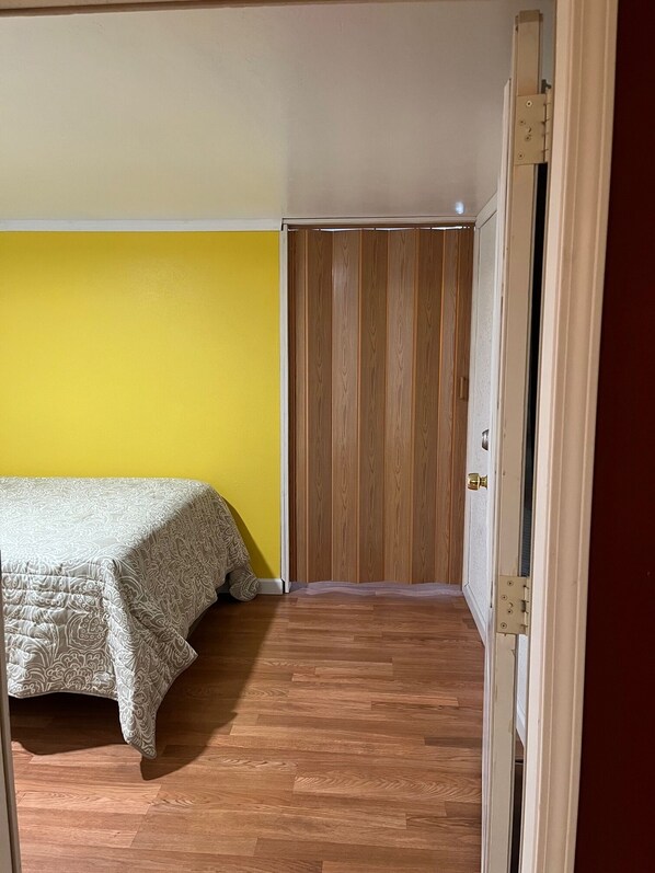A bedroom with a bed, a yellow wall, a wooden wardrobe, and a wooden floor.