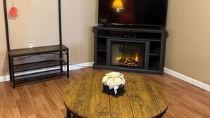 Un salón con televisor, chimenea, piso de madera y una mesa redonda con un ramo de flores.