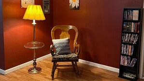 Una habitación con una silla de madera, una lámpara, una estantería y un cuadro floral en la pared.