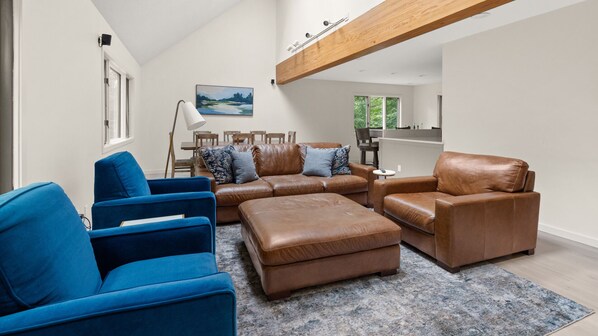A modern living room with a sectional sofa, a blue armchair, and a brown leather armchair. There is a dining area with wooden chairs and a table, and a large window providing a view of greenery.