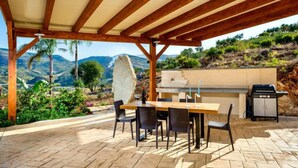 An outdoor patio with a wooden pergola, stone flooring, a dining table with chairs, a grill, and a view of a mountainous landscape. - Le Muse by Interhome (Castellammare del Golfo)