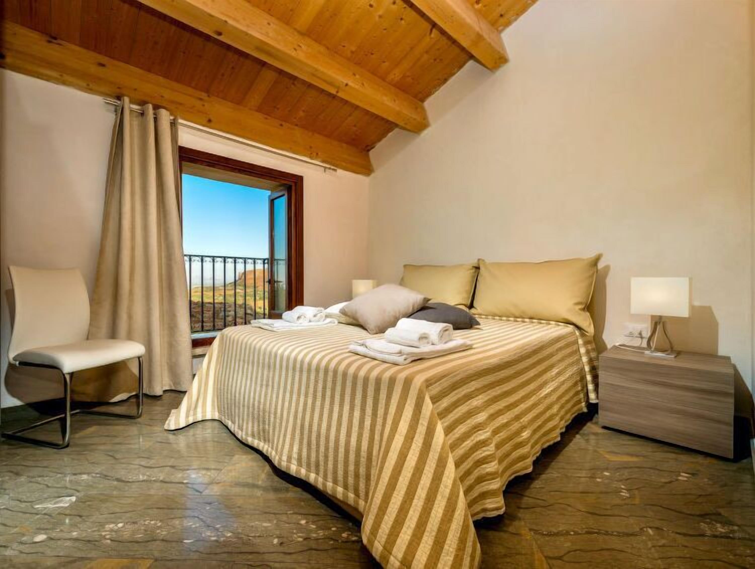A bedroom with a bed, a chair, a nightstand, a window with curtains, and a balcony with a view.