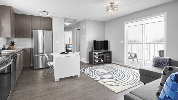 A modern kitchen with stainless steel appliances, a dining area with a white table and chairs, a grey sofa, and a large sliding glass door to a balcony. - Grey Havens Laneway home w/ garage, balcony, AC near WEM! (Edmonton)