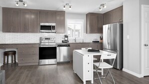A modern kitchen with dark wood cabinets, stainless steel appliances, and a white island with seating. - Grey Havens Laneway home w/ garage, balcony, AC near WEM! (Edmonton)