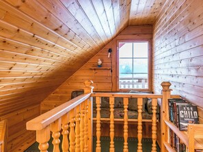 Un interior de madera con techo inclinado, una ventana con vistas, una escalera de balaustres de madera y una estantería con libros.