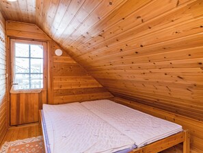 Een kamer met een schuin houten plafond, een enkel bed met matras, een raam met uitzicht op bomen en een deur.