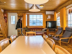 Interior de una cabaña de madera con una mesa de comedor, sillas, un sofá, un televisor y una ventana con vistas nevadas.