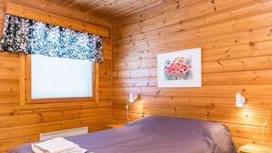 A wooden cabin room with a bed, a window with blinds, a framed picture on the wall, and a bedside table with a lamp.