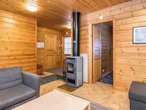 Een houten blokhutinterieur met een houtkachel in het midden, twee grijze banken en een kleine tafel.