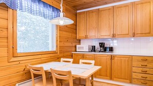A wooden cabin interior with a dining table, chairs, a window with blinds, a hanging light fixture, wooden cabinets, a microwave, and a coffee maker.