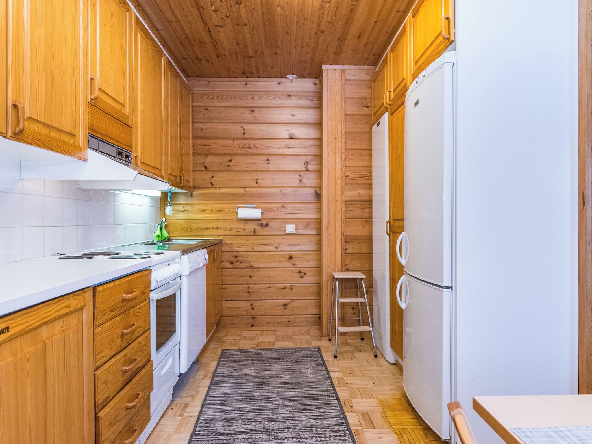 A compact kitchen with wooden cabinets, a white countertop, a built-in oven, a refrigerator, and a small stool.