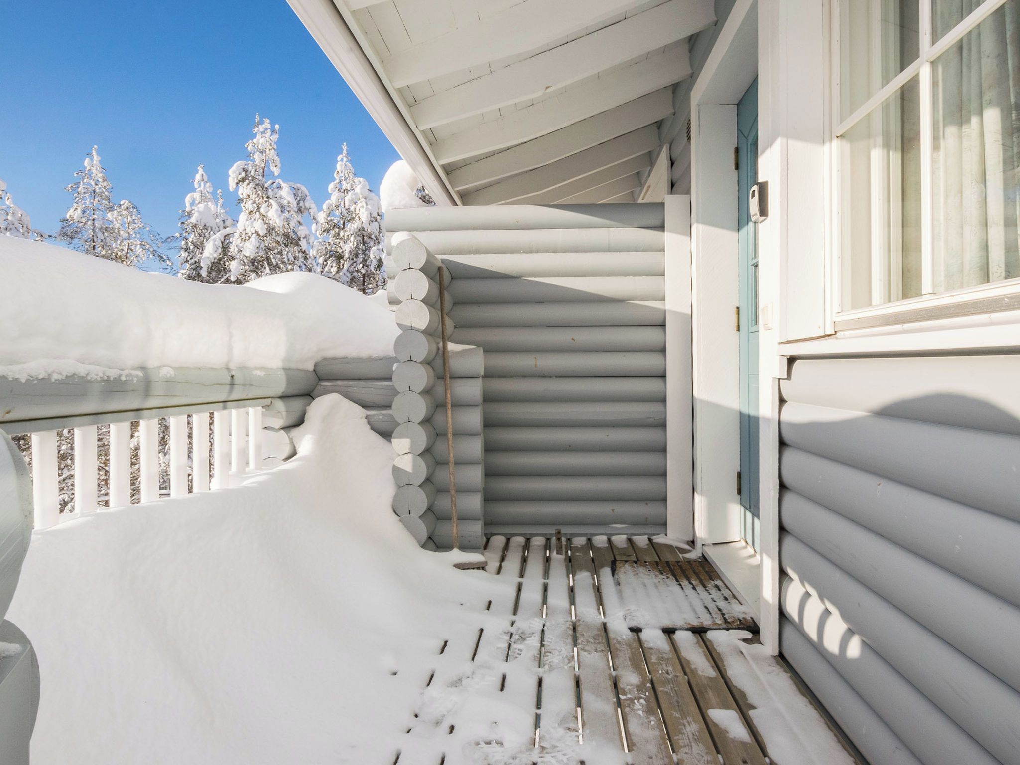 A snow-covered wooden deck with a metal railing, a stack of cylindrical logs, and a window with a blue trim.