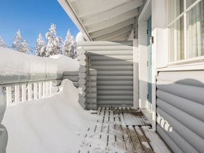 Une terrasse en bois recouverte de neige, avec une rambarde en métal, une pile de bûches de bois cylindriques et une fenêtre dotée d’un cadre bleu.
