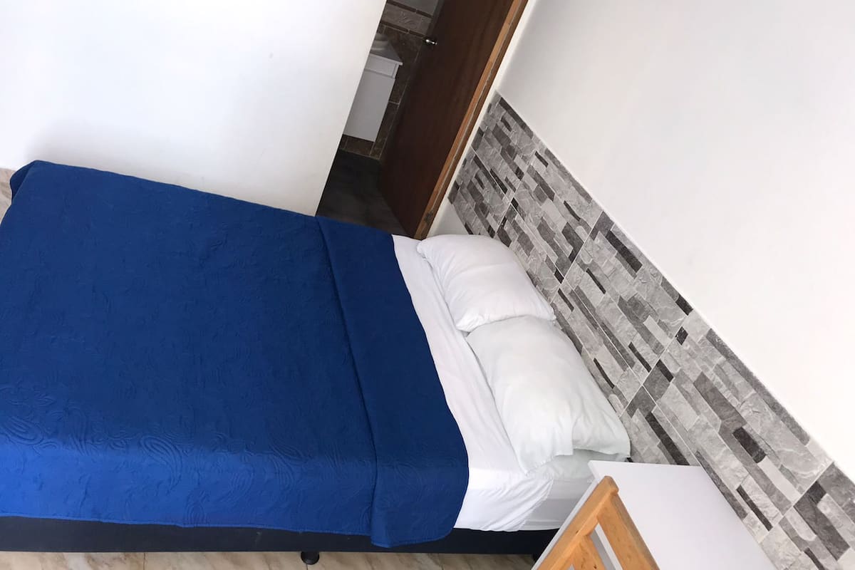 Ein Einzelbett mit blauer Bettdecke und weißen Kissen, ein hölzernes Kopfbrett mit grau-weißer Fliesenmusterung und ein kleiner Holzstuhl.