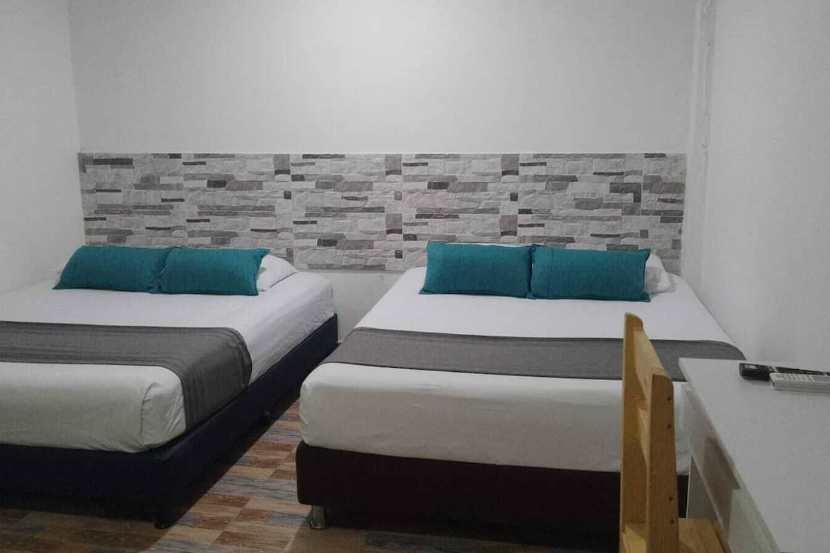 Ein Zimmer mit zwei Betten, jedes mit einem türkisblauen Kissen und einer weißen Decke. Das Zimmer hat eine Fliesenwand und einen Holzstuhl.