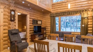 A log cabin interior with a dining area, a television, and a large window with a view of snow-covered trees.