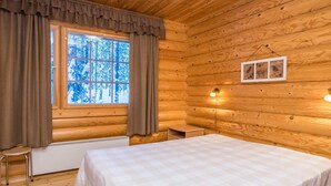 A log cabin bedroom with a bed, a window with curtains, a radiator, a small table, and a wall-mounted light fixture.