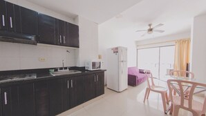 Cozinha compacta com armários pretos, micro-ondas e geladeira. Há uma área de refeições com mesa e cadeiras, e um ventilador de teto.