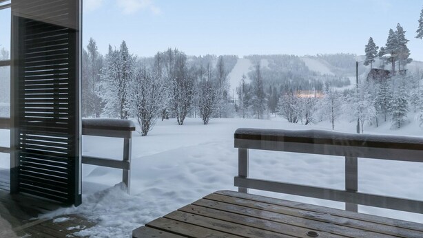A wooden bench and a window overlooking a snowy landscape with trees and a distant building.