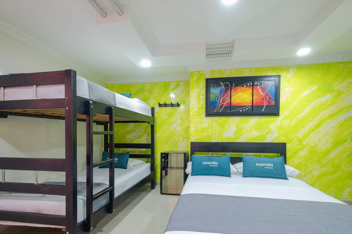 A hotel room with bunk beds, a bed with a grey blanket, a small bedside table, and a green wall with a framed picture.