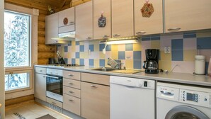 A compact kitchen with wooden cabinets, a dishwasher, a washing machine, and a window with a view of trees. - Hallantähti mökki by Interhome (Hyrynsalmi)